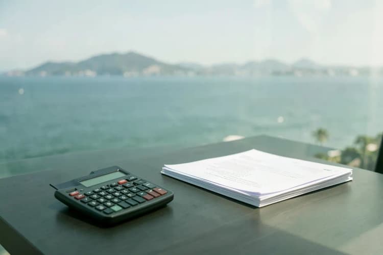 Calculadora e documentos sobre uma mesa com vista para o mar.